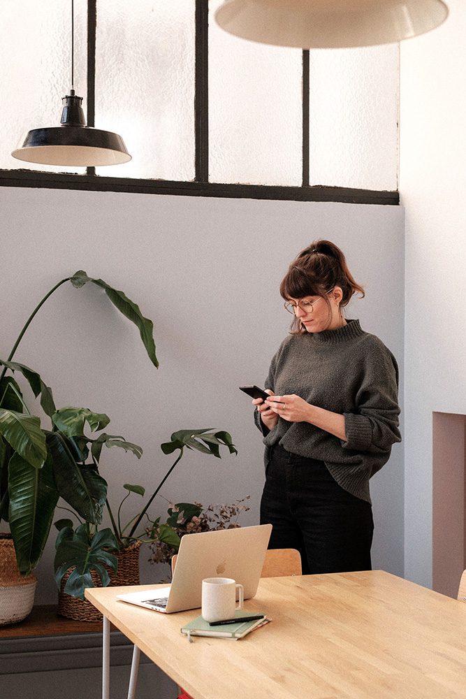offre-mentorat-dorothee-cadiot Dorothée Cadiot est debout en train de regarder son téléphone, à côté il y a une table sur laquelle on aperçoit un ordinateur, un cahier et un mug.